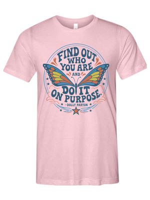 A light pink short-sleeve crewneck t-shirt featuring a colorful graphic print on the front. The graphic displays a butterfly with blue, yellow, and orange wings, surrounded by the motivational quote 'FIND OUT WHO YOU ARE AND DO IT ON PURPOSE' in blue lettering, with 'DOLLY PARTON' printed below.