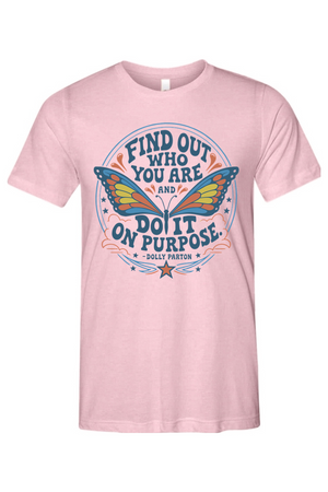 A light pink short-sleeve crewneck t-shirt featuring a colorful graphic print on the front. The graphic displays a butterfly with blue, yellow, and orange wings, surrounded by the motivational quote 'FIND OUT WHO YOU ARE AND DO IT ON PURPOSE' in blue lettering, with 'DOLLY PARTON' printed below.