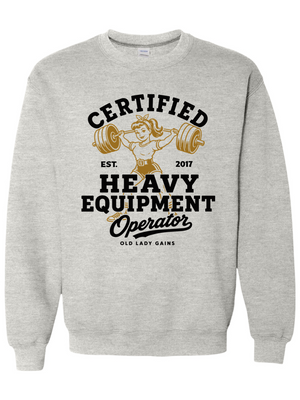 A light heather gray crewneck sweatshirt featuring a vintage-style graphic on the front with a cartoon illustration of an older woman lifting a barbell and the text 'CERTIFIED HEAVY EQUIPMENT Operator'.