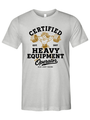 A light gray short-sleeve t-shirt with a vintage-style graphic on the front. The graphic features a retro illustration of a woman lifting a barbell, with the text 'CERTIFIED HEAVY EQUIPMENT Operator' in black and gold lettering.