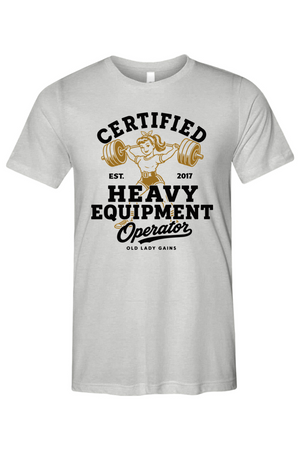 A light gray short-sleeve t-shirt with a vintage-style graphic on the front. The graphic features a retro illustration of a woman lifting a barbell, with the text 'CERTIFIED HEAVY EQUIPMENT Operator' in black and gold lettering.