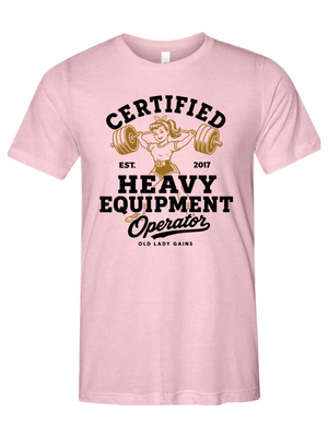 A light pink short-sleeve crewneck t-shirt featuring a vintage-style graphic on the front with the text 'CERTIFIED HEAVY EQUIPMENT Operator' and an illustration of a woman lifting a barbell.