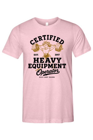 A light pink short-sleeve crewneck t-shirt featuring a vintage-style graphic on the front with the text 'CERTIFIED HEAVY EQUIPMENT Operator' and an illustration of a woman lifting a barbell.