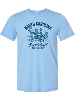 Light blue t-shirt with a graphic design of a person in a plane holding a pickleball paddle and ball, with text 'North Carolina Pickleball Takes Flight' on a white background.