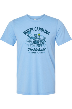 Light blue t-shirt with a graphic design of a person in a plane holding a pickleball paddle and ball, with text 'North Carolina Pickleball Takes Flight' on a white background.