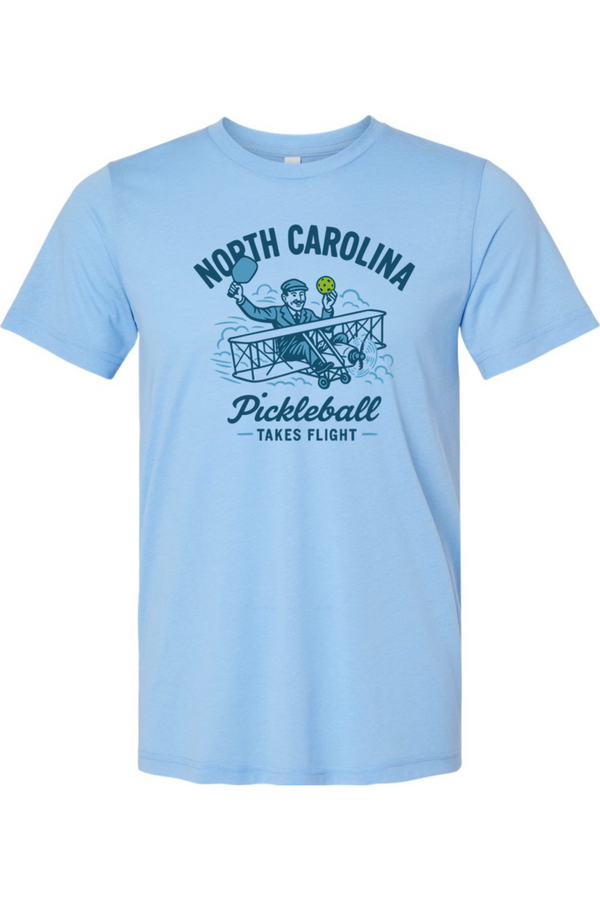 Light blue t-shirt with a graphic design of a person in a plane holding a pickleball paddle and ball, with text 'North Carolina Pickleball Takes Flight' on a white background.