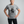An older woman with short gray hair stands against a light gray studio background wearing a heather gray graphic t-shirt with a black cat illustration and the text 'It's Fine. I'm Fine.