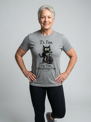 An older woman with short gray hair stands against a light gray studio background wearing a heather gray graphic t-shirt with a black cat illustration and the text 'It's Fine. I'm Fine.
