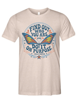 A light beige crewneck t-shirt featuring a colorful retro-style graphic with a butterfly and the quote 'FIND OUT WHO YOU ARE AND DO IT ON PURPOSE' by Dolly Pardon