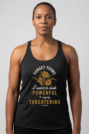 A woman wearing a dark heather gray racerback tank top with a gold and white graphic print that reads 'FORGET YOUNG I want to look POWERFUL & VAGUELY THREATENING' with a snake illustration. The brand 'Old Lady Gains' is printed at the bottom of the graphic.