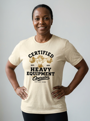 A smiling woman wearing a cream-colored graphic t-shirt with a black and gold print that reads 'CERTIFIED HEAVY EQUIPMENT Operator' with an illustration of a woman lifting weights. She is posing with her hands on her hips against a solid light gray background.