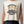 A woman wearing a sleeveless cream-colored muscle tank top with a vintage-style graphic that reads 'CERTIFIED HEAVY EQUIPMENT Operator' in black and gold, paired with dark patterned leggings against a solid gray background.