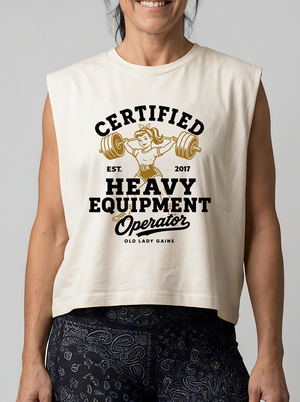 A woman wearing a sleeveless cream-colored muscle tank top with a vintage-style graphic that reads 'CERTIFIED HEAVY EQUIPMENT Operator' in black and gold, paired with dark patterned leggings against a solid gray background.