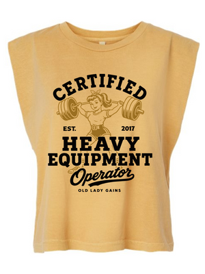 A sleeveless, cropped t-shirt in a warm mustard yellow color, featuring a vintage-style black graphic print on the front with the text 'CERTIFIED HEAVY EQUIPMENT Operator' and an illustration of an older woman lifting a barbell.