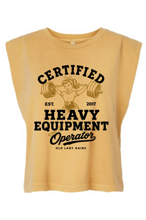 A sleeveless, cropped t-shirt in a warm mustard yellow color, featuring a vintage-style black graphic print on the front with the text 'CERTIFIED HEAVY EQUIPMENT Operator' and an illustration of an older woman lifting a barbell.