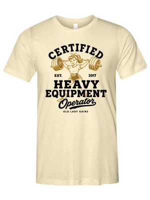 A cream-colored short-sleeve t-shirt with a vintage-style graphic on the front. The graphic features a retro illustration of a woman lifting a barbell, surrounded by bold black text that reads 'CERTIFIED HEAVY EQUIPMENT Operator' and smaller text 'EST.