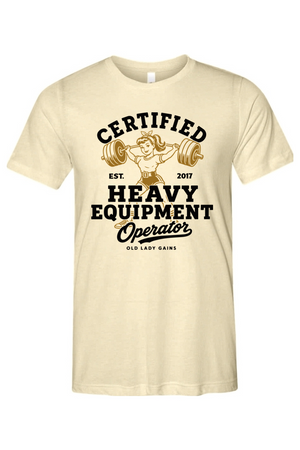 A cream-colored short-sleeve t-shirt with a vintage-style graphic on the front. The graphic features a retro illustration of a woman lifting a barbell, surrounded by bold black text that reads 'CERTIFIED HEAVY EQUIPMENT Operator' and smaller text 'EST.