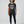 A woman with curly gray hair stands against a light gray studio background wearing a dark charcoal heathered t-shirt with the text 'LIFTING IS FAVORITE SPORT' in white and a thin pink stripe, paired with black leggings and black sneakers.