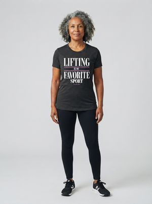 A woman with curly gray hair stands against a light gray studio background wearing a dark charcoal heathered t-shirt with the text 'LIFTING IS FAVORITE SPORT' in white and a thin pink stripe, paired with black leggings and black sneakers.