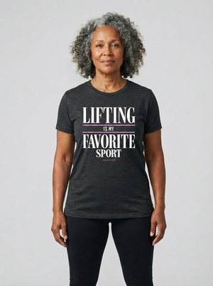 A woman with curly gray hair wearing a dark heather gray graphic t-shirt that reads 'LIFTING IS MY FAVORITE SPORT' in white and purple text, paired with black leggings, against a light gray studio background.