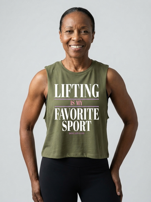 A smiling woman wearing an olive green sleeveless graphic tank top with the text 'LIFTING IS MY FAVORITE SPORT' in white and pink, paired with black leggings, against a solid light gray background.