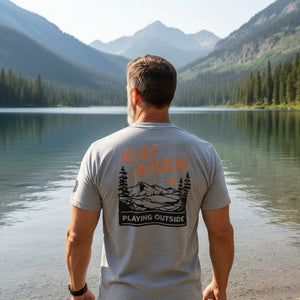 Man wearing a t-shirt that say Never Outgrow Playing Outside standing by a lake and mountains