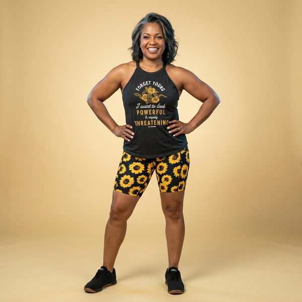 Woman wearing a black Old Lady Gains Powerful & Vaguely Threatening tank top with text and CVG sunflower shorts on a Gold background