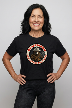 A muscular, active middle-aged woman wearing a heather charcoal black triblend unisex t-shirt. The graphic features a retro, circular ranger bear design pointing forward and holding a barbell, with bold red and cream text that reads: REMEMBER Only You Can Age Strong.