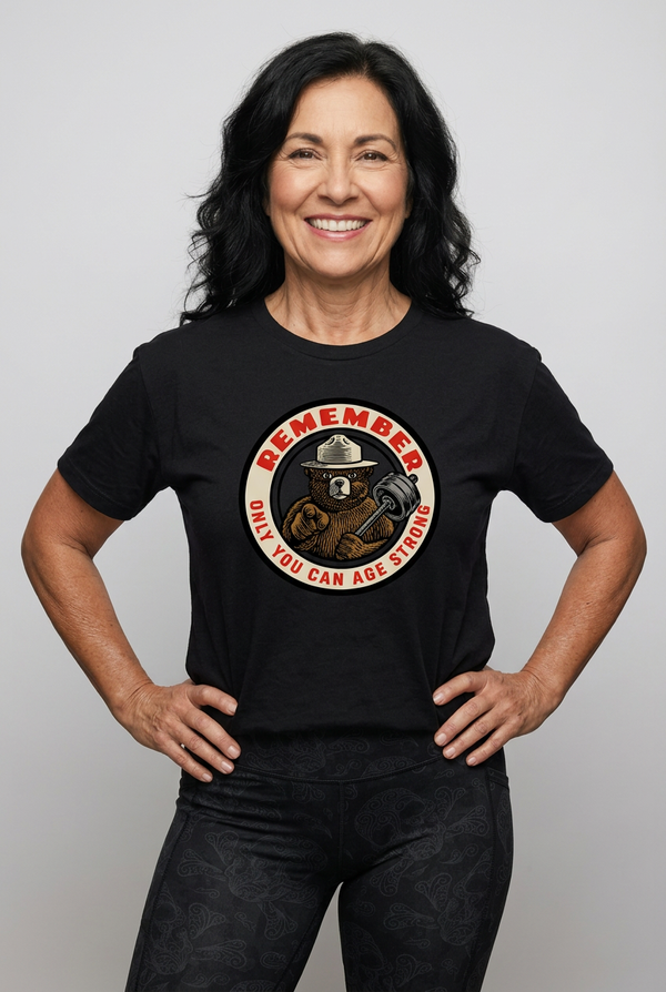 A muscular, active middle-aged woman wearing a heather charcoal black triblend unisex t-shirt. The graphic features a retro, circular ranger bear design pointing forward and holding a barbell, with bold red and cream text that reads: REMEMBER Only You Can Age Strong.