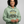 A smiling woman with curly black hair wearing a sage green pullover hoodie with a graphic that reads 'CERTIFIED HEAVY EQUIPMENT Operator' in black and gold, posed against a solid gray studio background.