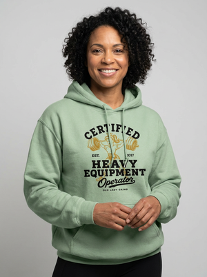 A smiling woman with curly black hair wearing a sage green pullover hoodie with a graphic that reads 'CERTIFIED HEAVY EQUIPMENT Operator' in black and gold, posed against a solid gray studio background.