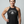 A woman modeling a dark charcoal gray heathered tank top with a white graphic that reads 'DEADLIFT' above a skull and crossbones, set against a solid light gray background.