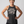 A smiling woman with her hands on her hips wears a dark heather gray racerback tank top with a white printed quote that reads 'women grow more radical with age -florida giraffe'. She is wearing black leggings against a solid light gray studio background.