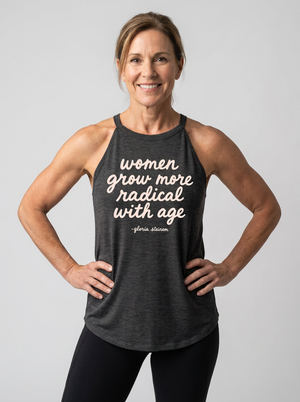 A smiling woman with her hands on her hips wears a dark heather gray racerback tank top with a white printed quote that reads 'women grow more radical with age -florida giraffe'. She is wearing black leggings against a solid light gray studio background.