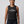 A smiling woman with braided hair stands against a light gray studio background wearing a dark charcoal sleeveless tank top with white text reading 'AGING Disgracefully' and 'OLD LADY CLINS,' paired with dark leggings.