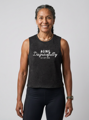 A smiling woman with braided hair stands against a light gray studio background wearing a dark charcoal sleeveless tank top with white text reading 'AGING Disgracefully' and 'OLD LADY CLINS,' paired with dark leggings.