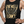 Woman over 40 wearing a black tank top with gold graphic and text against a plain background