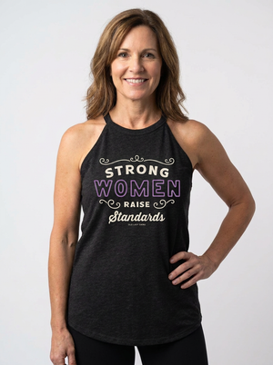 A smiling woman wearing a dark charcoal heather halter tank top with the text 'STRONG WOMEN RAISE Standards' printed on the front, posed against a light gray studio background.
