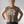 A woman wearing a sleeveless taupe-brown crop top with a white printed quote that reads 'women grow more radical with age -gloria atenera', paired with dark paisley leggings, standing against a light gray background.