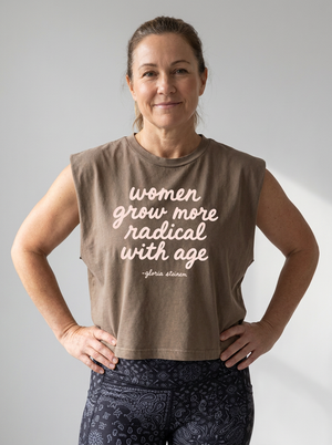 A woman wearing a sleeveless taupe-brown crop top with a white printed quote that reads 'women grow more radical with age -gloria atenera', paired with dark paisley leggings, standing against a light gray background.