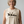 A smiling woman with short gray hair wearing a beige sleeveless crop top with a black graphic of horses and riders and the text 'RAISED UNGOVERNABLE. RAISING UNGOVERNABLE.