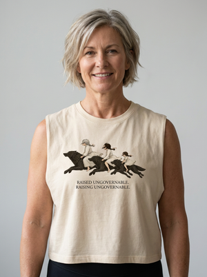 A smiling woman with short gray hair wearing a beige sleeveless crop top with a black graphic of horses and riders and the text 'RAISED UNGOVERNABLE. RAISING UNGOVERNABLE.