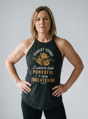 A woman modeling a dark charcoal gray racerback tank top with a gold graphic print that reads 'FORGET YOUNG I want to look POWERFUL & vaguley THREATENING'.