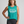 A woman modeling a teal sleeveless muscle tank top with the text 'Someone's Badass Grandma' printed in white on the front, paired with dark gray leggings, against a light gray studio background.