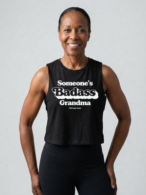 A smiling woman wearing a black sleeveless graphic tank top with the text 'Someone's Badass Grandma' in white, paired with black leggings, against a light gray studio background.