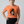 A woman modeling a heather orange crewneck t-shirt with a graphic of a raven holding a sign that reads 'IS HE YET?'. She is wearing black leggings against a solid light gray background.