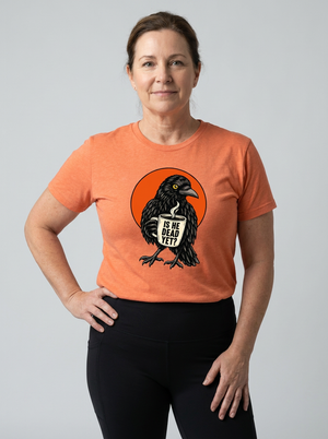 A woman modeling a heather orange crewneck t-shirt with a graphic of a raven holding a sign that reads 'IS HE YET?'. She is wearing black leggings against a solid light gray background.