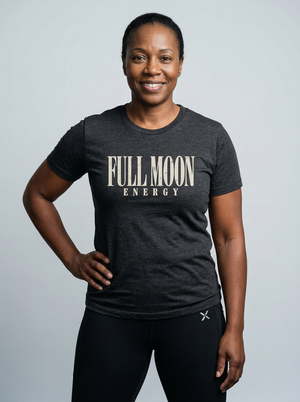 A smiling woman wearing a dark heather gray t-shirt with 'FULL MOON ENERGY' printed in beige on the chest, paired with black athletic leggings, against a light gray studio background.