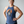 A smiling woman wearing a heathered blue racerback tank top with a graphic of a raven in front of an orange circle and the text 'IS HE DEAD YET?' on the front, paired with black leggings, standing against a light gray studio background.
