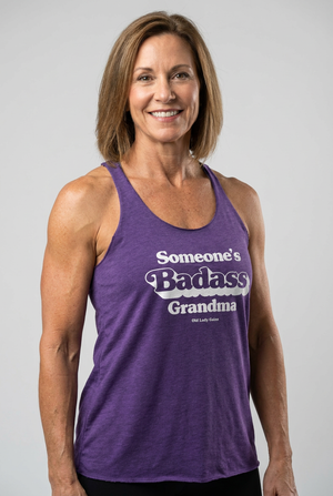 A smiling woman with shoulder-length brown hair wearing a purple heathered racerback tank top with the text 'Someone's Badass Grandma' in white and gray, photographed against a light gray studio background.